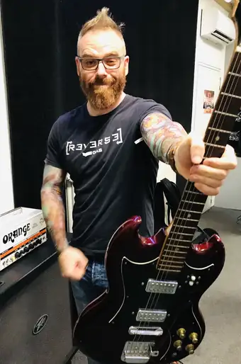 Klein Péter próbateremben, elektromos gitárt tart a nyakánál fogva a kamera felé; mögötte Orange erősítőfej és hangláda.